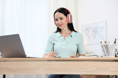 Kulaklıklı bir kadın, bilgisayarla kapalı bir masada çevrimiçi sınavdan geçerken not alıyor.