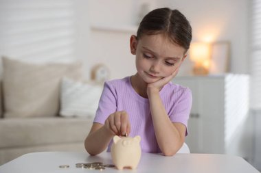 Küçük kız, içerideki beyaz masadaki kumbaraya cep harçlığı koyuyor.