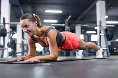 Gülümseyen kadın spor salonunda yoga minderinde kalas egzersizi yapıyor.