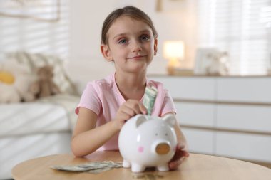 Küçük kız, ahşap masadaki domuz kumbarasına cep harçlığı koyuyor.