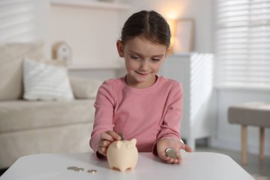 Küçük kız, içerideki beyaz masadaki kumbaraya cep harçlığı koyuyor.