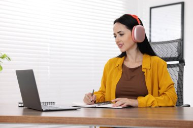 Kulaklıklı bir kadın, bilgisayarla kapalı bir masada çevrimiçi sınavdan geçerken not alıyor.