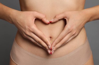 Düz göbekli genç bir kadın gri arka planda kalp hareketi yapıyor, yakın plan.