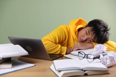 Sınavdan önce yeşil duvarın yanındaki tahta masada dizüstü bilgisayarı ve kitapları olan yorgun bir öğrenci.
