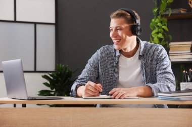 Kulaklıklı bir adam, içerideki masada dizüstü bilgisayarla sınavdan geçerken not alıyor.