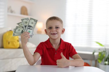 Küçük çocuk cep harçlığı tutuyor ve beyaz masada baş parmak gösteriyor.