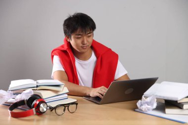 Dizüstü bilgisayarı ve kitapları olan öğrenci gri duvarın yanındaki tahta masada sınava hazırlanıyor.