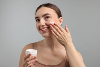 Gri arka planda yüzüne krem süren güzel bir kadın.
