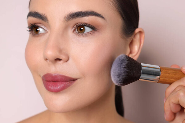Beautiful woman applying makeup with brush on beige background, closeup