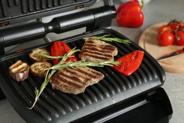 Open electric grill with beef steaks, rosemary, bell pepper and eggplant on light grey table, closeup