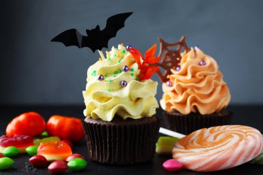 Yarasa soslu lezzetli kekler, çikolata parçaları, çikolata ağı ve gri arka planda koyu dokulu masa üzerinde renkli şekerler, yakın plan. Cadılar Bayramı