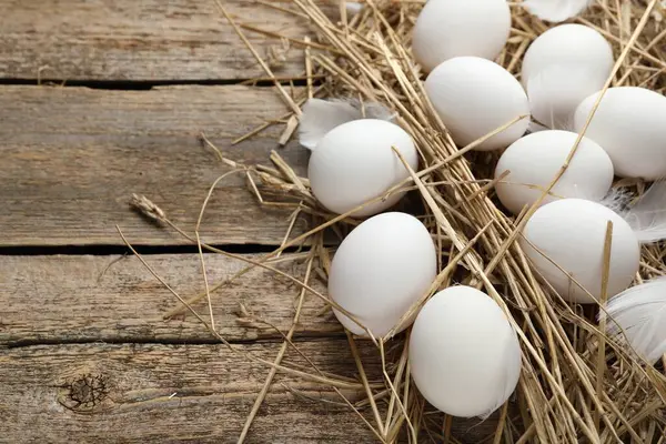 Raw chicken eggs, feathers and straw on wooden table, closeup. Space for text