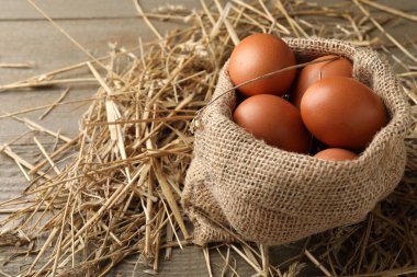 Raw chicken eggs in burlap sack and straw on wooden table, closeup. Space for text