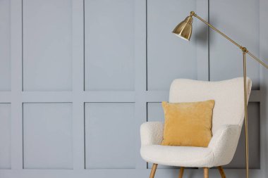 Stylish lamp and comfortable armchair with cushion near gray wall indoors. Space for text
