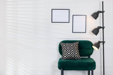 Stylish lamp, comfortable armchair with cushion and picture frames on white wall indoors. Space for text