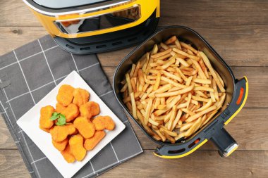Air fryer with cooked fries and chicken nuggets on wooden table, flat lay