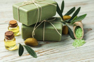 Natural soap bars with olives, sea salt, oil and green leaves on white wooden table, closeup