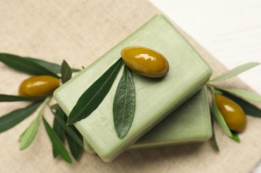 Natural soap bars with olives and green leaves on white table, closeup