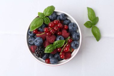 Different ripe berries and basil leaves in bowl on white tiled table, flat lay
