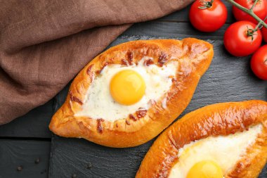 Tasty khachapuri with cheese, eggs and tomatoes on black wooden table, flat lay