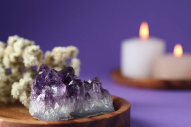 Beautiful amethyst gemstone, flowers and burning candles on purple background, closeup. Space for text