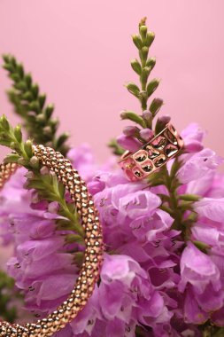 Pembe arka planda zarif altın yüzük, bilezik ve güzel çiçekler, yakın plan.