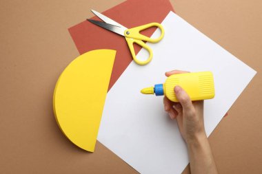 Kadın, kahverengi masada yapıştırıcı ve renkli kağıtlarla el işi yapıyor.