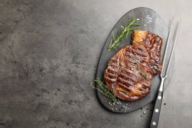 Delicious grilled beef steak with herbs, spices and carving fork on grey textured table, top view. Space for text