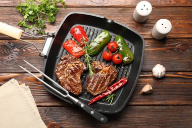 Pan with grilled beef steaks, rosemary, vegetables, shakers and carving fork on wooden table, flat lay