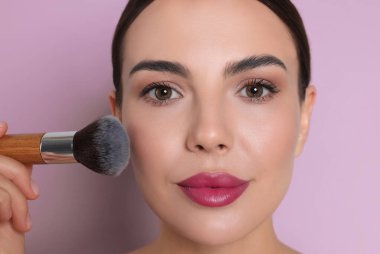 Leylak rengi arka planda fırçayla makyaj yapan güzel kadın, yakın plan.