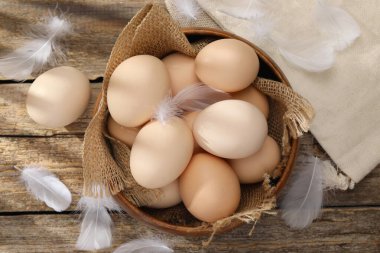 Raw chicken eggs in bowl and feathers on wooden table, top view