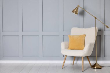 Stylish lamp and comfortable armchair with cushion near gray wall indoors. Space for text