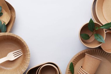 Set of disposable tableware and green leaves on white background, flat lay. Space for text