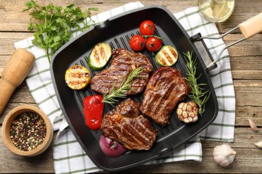 Pan with grilled beef steaks, rosemary, spices and vegetables on wooden table, flat lay