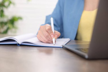 İçerideki ahşap masada dizüstü bilgisayardan geçerken not alan kadın, yakın plan.