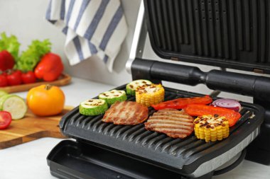Electric grill with tasty meat and vegetables on white table in kitchen, closeup