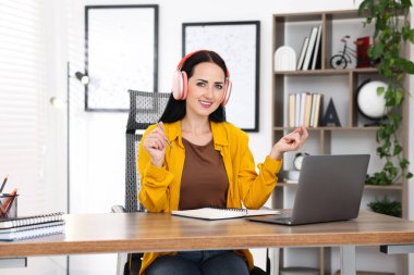 Kulaklıklı bir kadın bilgisayarla kapalı bir masada çevrimiçi sınavdan geçiyor.