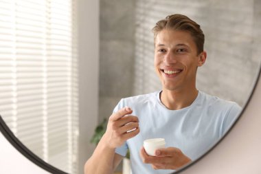 Evde aynanın yanında yüz kremi dolu kavanozu olan yakışıklı bir genç.