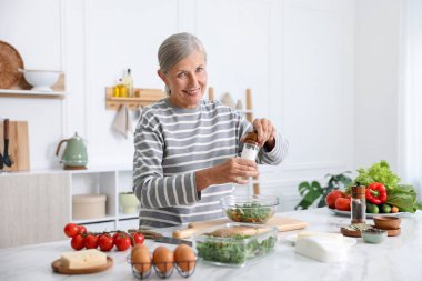 Yemek pişirme süreci. Üst düzey kadın mutfaktaki beyaz mermer masada salataya baharat ekliyor.