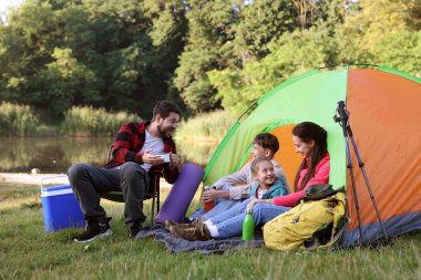 Mutlu bir aile kamp çadırı, turistik malzemeler ve nehir kenarında içki molası veriyor.