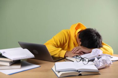 Sınavdan önce yeşil duvarın yanındaki tahta masada dizüstü bilgisayarı ve kitapları olan yorgun bir öğrenci.