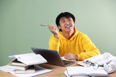 Laptoplu ve kitaplı mutlu öğrenci yeşil duvarın yanındaki tahta masada sınava hazırlanıyor.