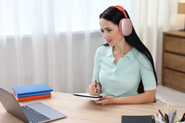 Kulaklıklı bir kadın, bilgisayarla kapalı bir masada çevrimiçi sınavdan geçerken not alıyor.