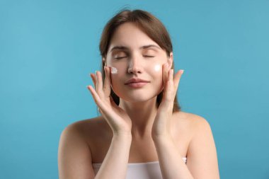 Güzel bir kadın açık mavi arka planda yüzüne krem sürüyor.