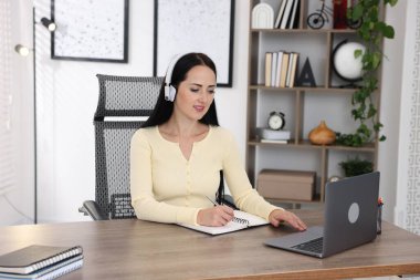 Kulaklıklı bir kadın bilgisayarla kapalı bir masada çevrimiçi sınavdan geçiyor.