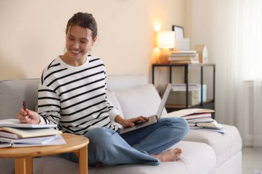 Bilgisayarlı, defterli ve kalemli gülümseyen öğrenci evdeki kanepede sınava hazırlanıyor.