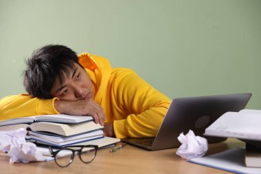 Sınavdan önce yeşil duvarın yanındaki tahta masada dizüstü bilgisayarı ve kitapları olan yorgun bir öğrenci.