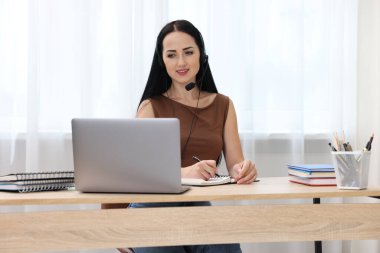 İçerideki masada dizüstü bilgisayarla sınavdan geçerken notlar alan kulaklıklı bir kadın.