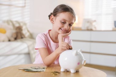 Küçük kız, ahşap masadaki domuz kumbarasına cep harçlığı koyuyor.