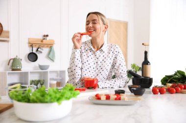 Mutfakta yemek pişirirken biber yiyen gülümseyen kadın.